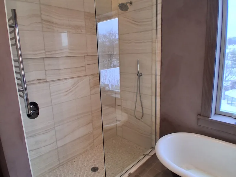 Walk-in shower with large stone-look wall tile, a frameless glass panel and a handheld shower, next to a clawfoot tub